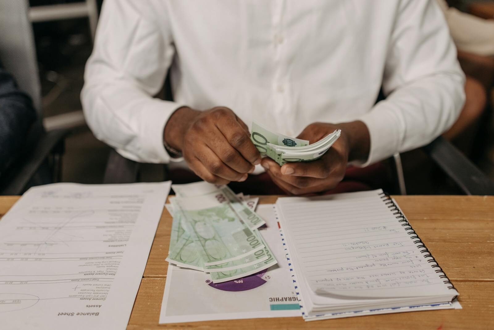 African businessman counting euro bills at a desk with documents and a notebook, highlighting finance theme.