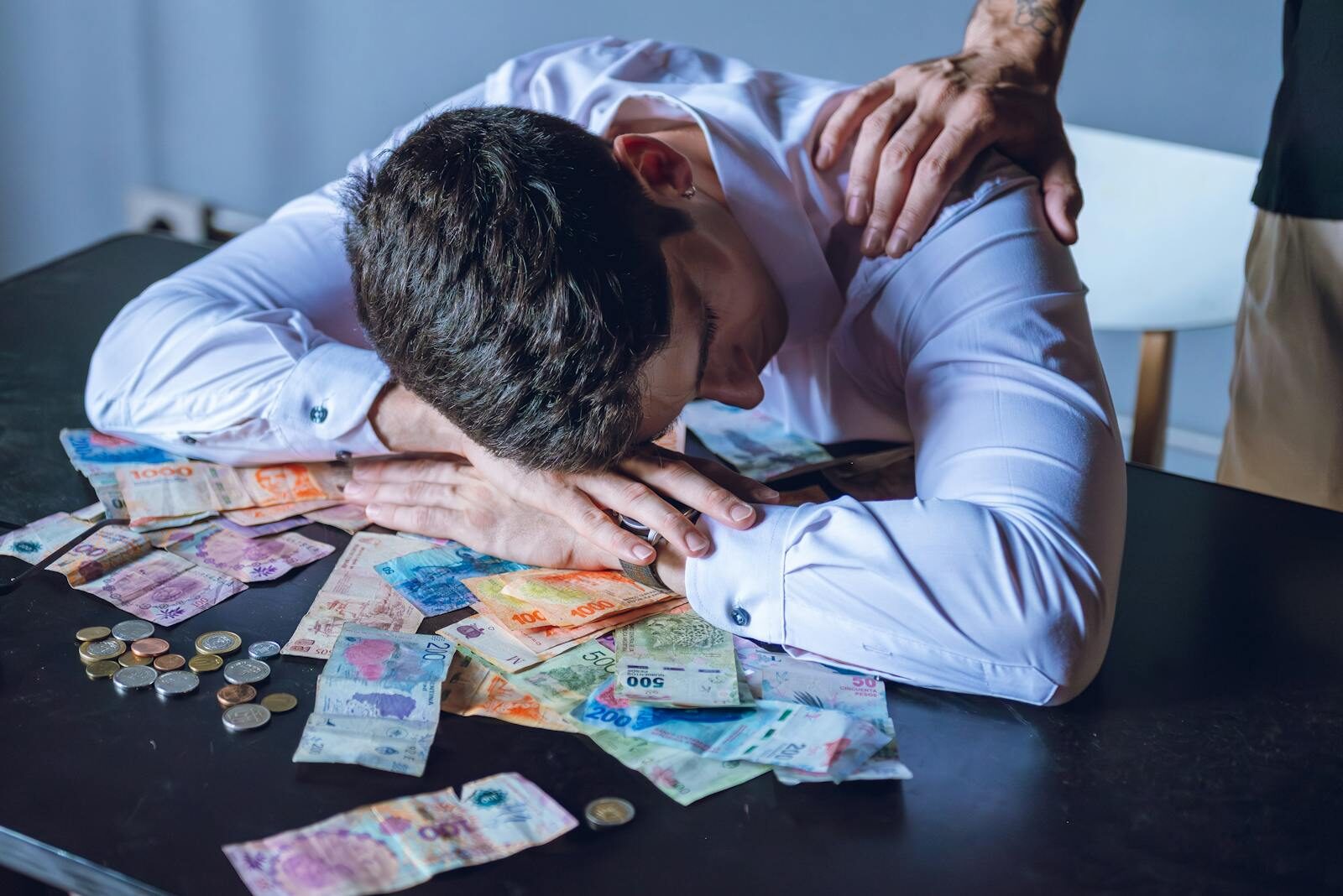 A man is overwhelmed with Argentine pesos, reflecting financial stress and support from another person.
