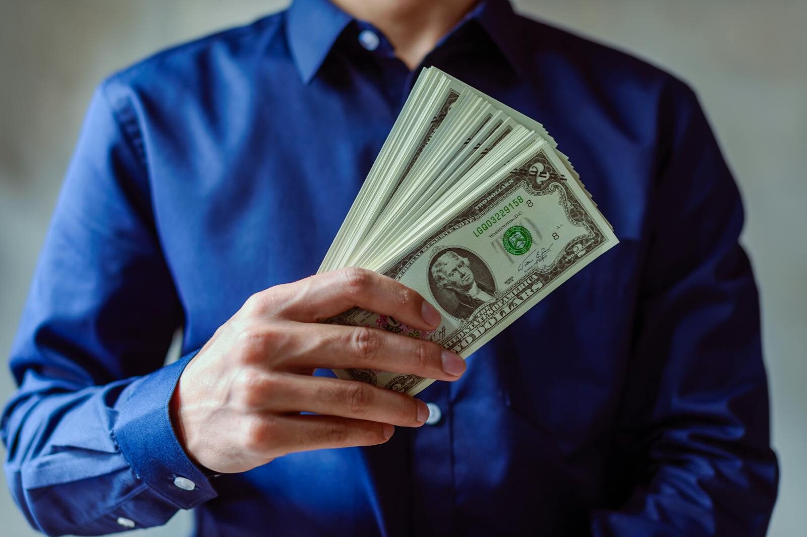 Close-up of a person in blue shirt holding multiple US dollar bills.
