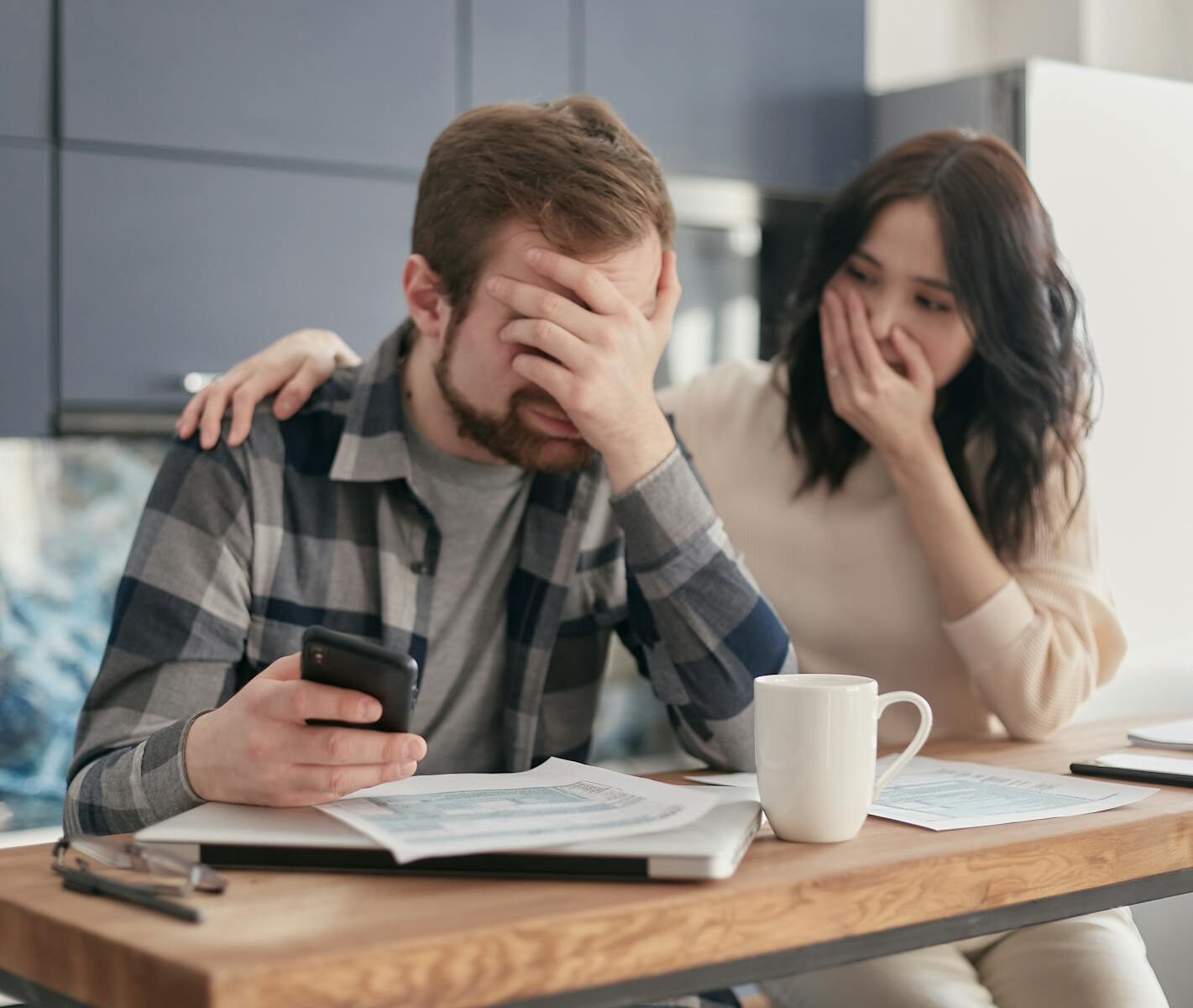 A couple looking stressed and holding a cellphone while reviewing financial documents indoors. Hands on faces in concern.