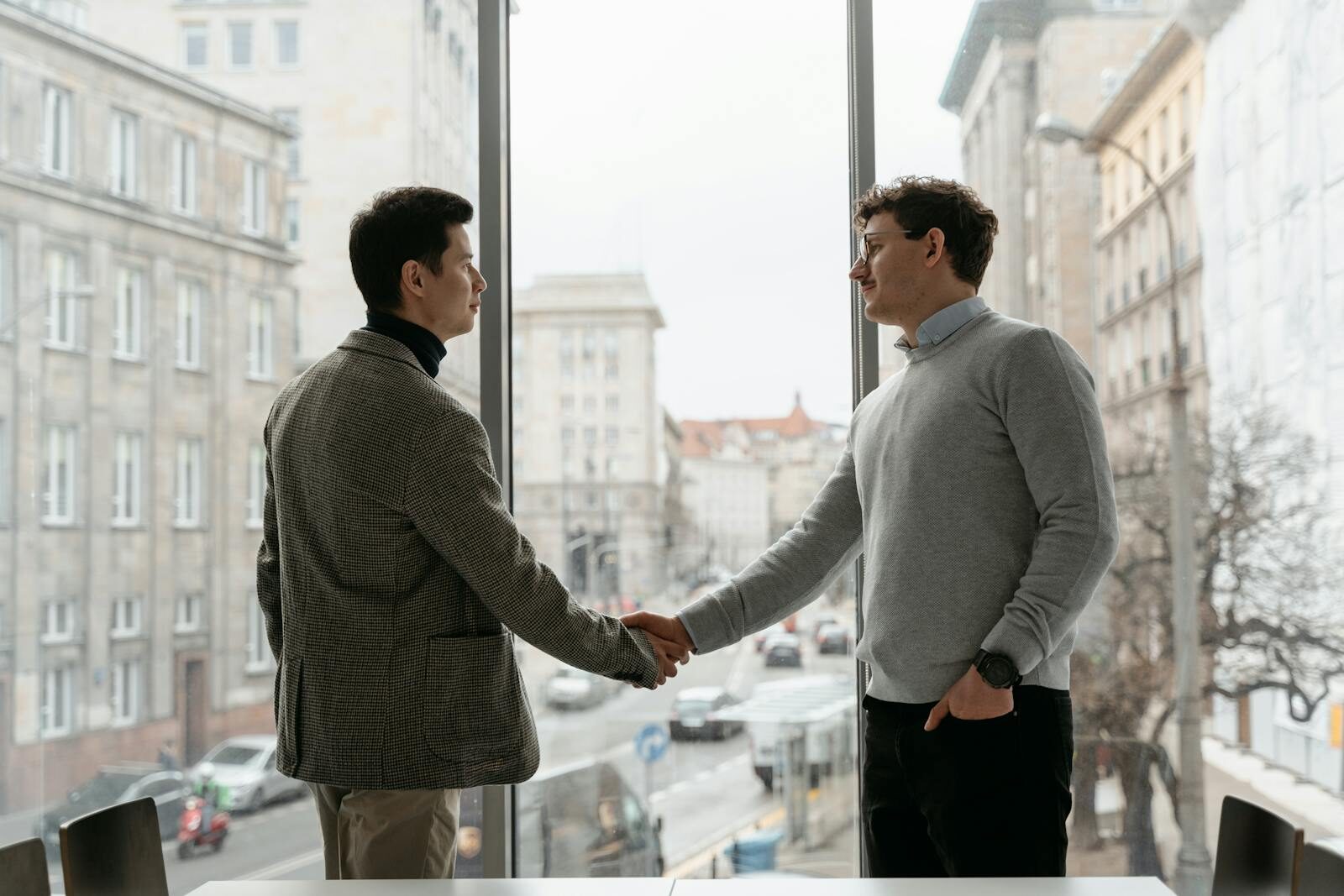 Two businessmen shaking hands in an urban office with city view through large windows.